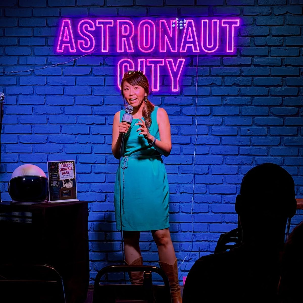 Dr. Ji performing stand-up with a microphone in front of a blue brick wall and a neon sign